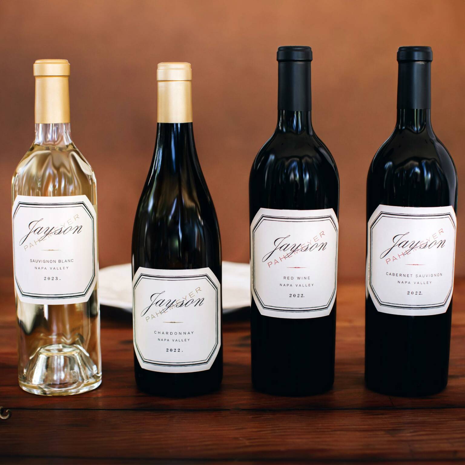 Four Jayson wine bottles are displayed on a wooden surface: a Sauvignon Blanc, a Chardonnay, a Red Wine, and a Cabernet Sauvignon, all labeled 2022 from Napa Valley.