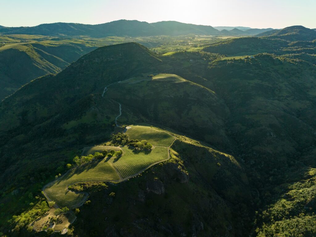 Aerial view of a lush, green hilly landscape with winding roads and valleys under a clear sky.