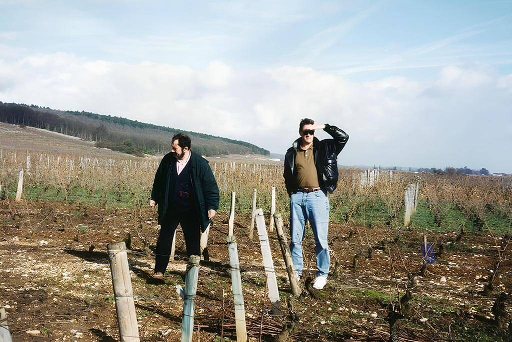 Jayson Pahlmeyer and Michel Rolland standing in a vineyard. One person is dressed in a dark coat, and the other in a leather jacket and jeans, shading their eyes with their hand. The vineyard is expansive under a blue sky.