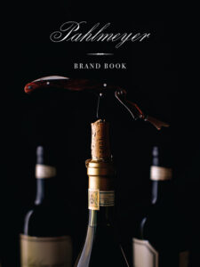 Close-up of a corkscrew opening a wine bottle, with the text "Pahlmeyer Brand Book" on a black background above. Blurred wine bottles are visible in the background.