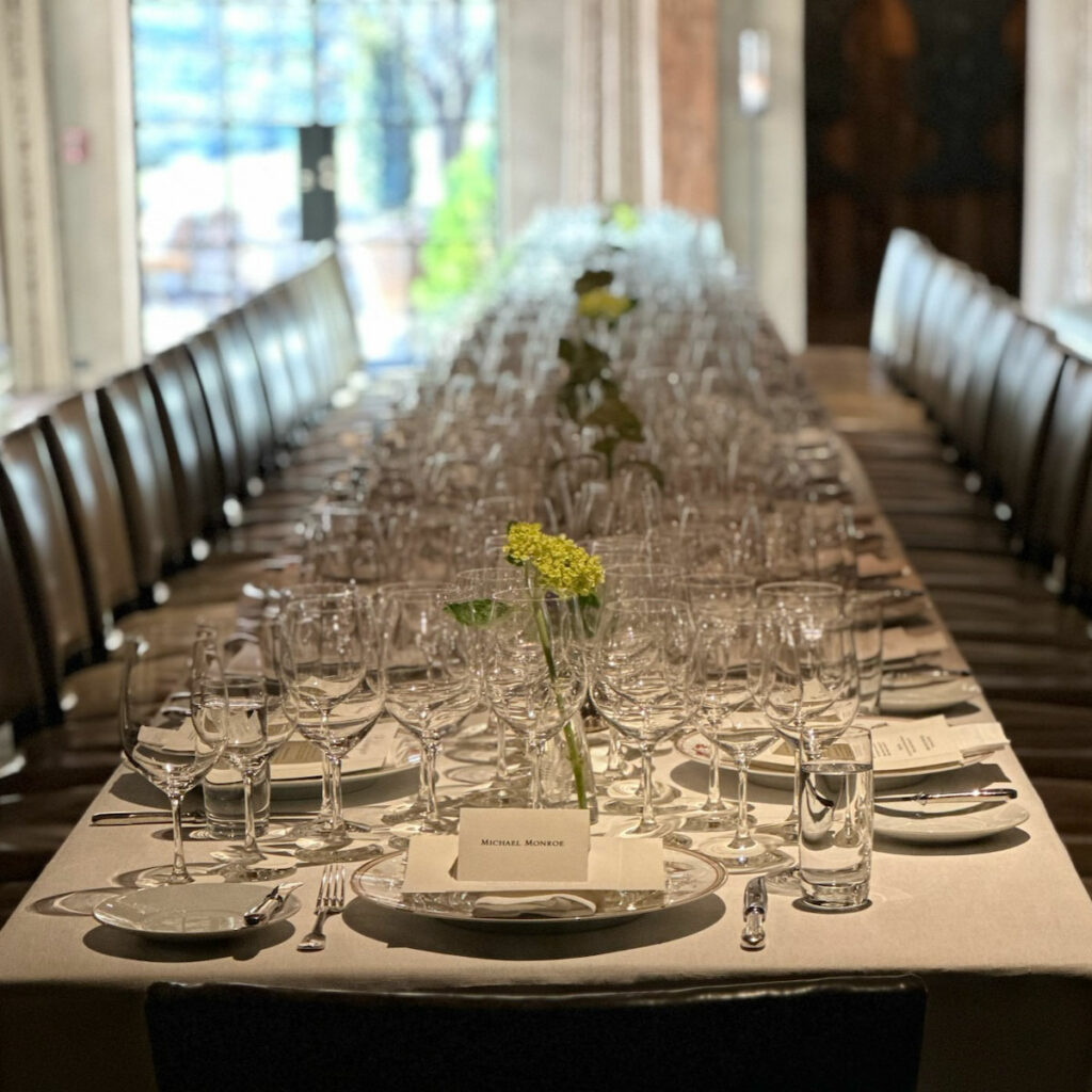 A long dining table is set with multiple place settings, including glassware and plates, and features a few floral centerpieces down the middle. The table is surrounded by chairs in a dimly lit room.