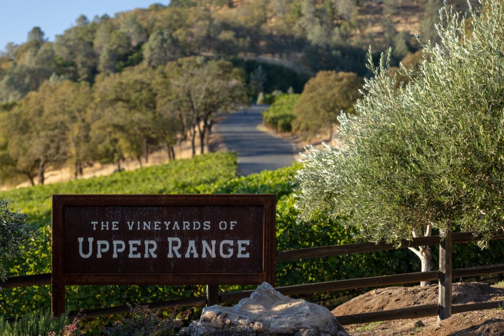 A sign reading "The Vineyards of Upper Range" stands in front of a vineyard with rows of grapevines and trees on a gently sloping hill.